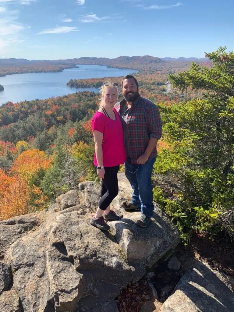 A man in a flannel shirt and jeans and a woman in a pink shirt stand close together, smiling. They are standing on a rocky hill, and behind them are hills, a river, and forests with fall colors.