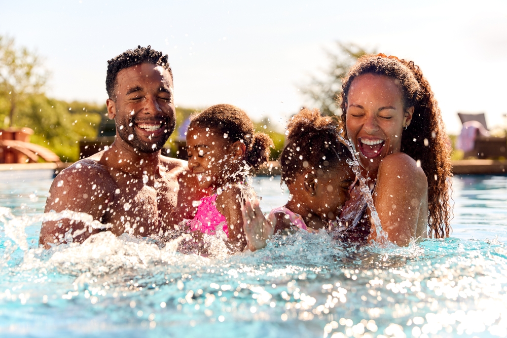 A family—a man and a woman and two young girls—are swimming in a pool. All are smiling, and water droplets are splashing around them.
