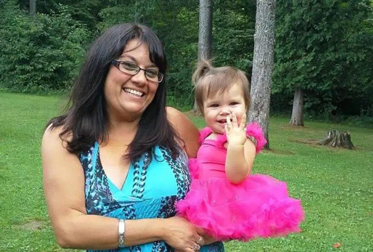 Image Alt tag: A smiling woman with brown hair and a blue dress holds a young girl in a pink tutu. Behind them is a grassy lawn and woods.