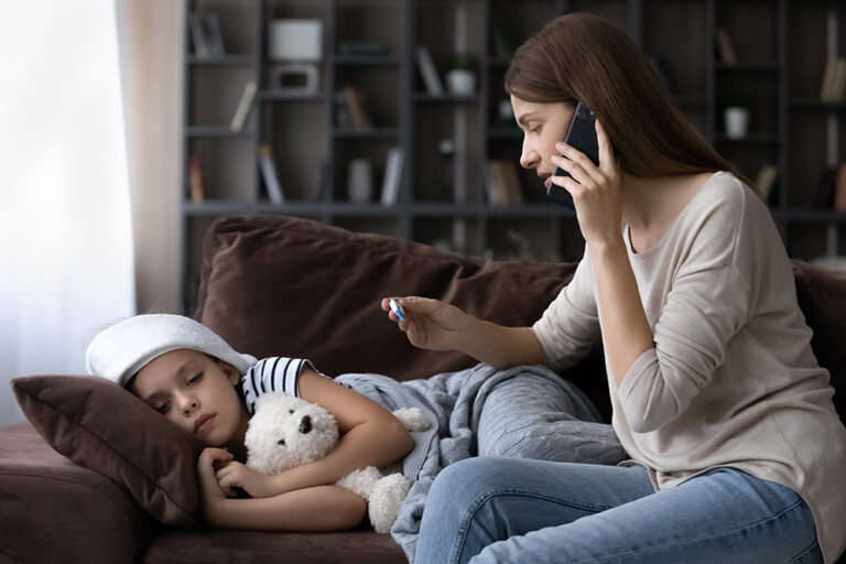 Image Alt tag: A girl lays on a couch with a washcloth on her forehead. A woman sits beside her, looking at a thermometer and talking on a cell phone.