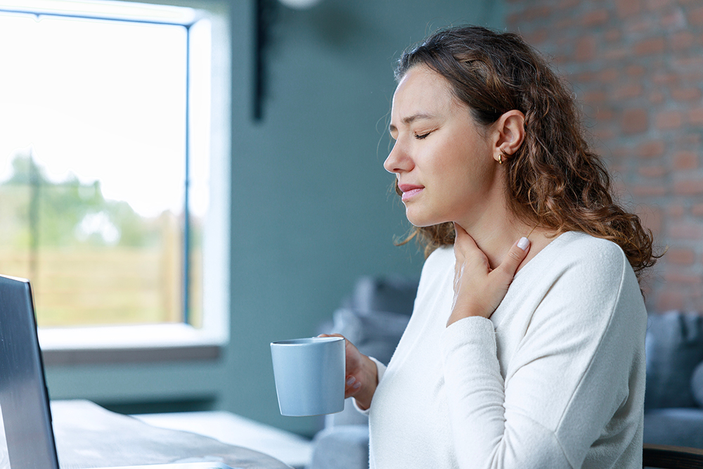 Image Alt tag:	A woman in a white shirt with brown hair sits at a laptop computer. She holds a cup and is touching her throat. Her eyes are closed in apparent pain.