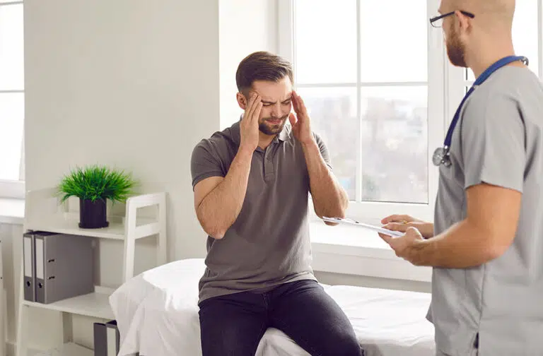 A male patient with his hands on his head in pain is consulting a doctor during a medical visit.