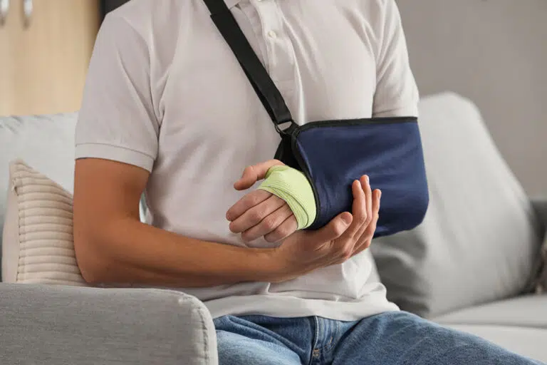 An injured young man sits on a sofa and holds his slung and casted arm.